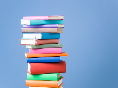 Multi colored books on blue background.The stack of books is on the left side of horizontal frame.Shot in studio with medium format camera.No people are seen in frame.